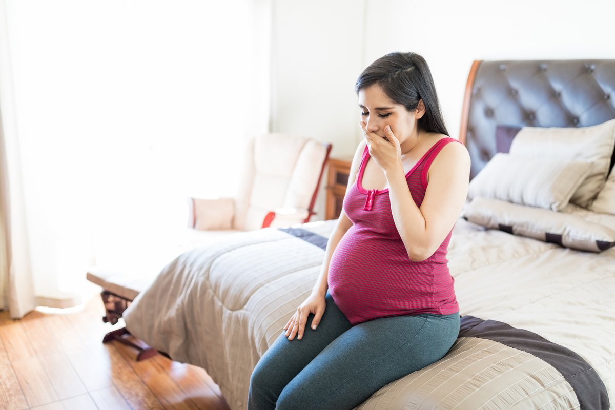 młoda kobieta w zaawansowanej ciąży siedzi na skraju łóżka i zakrywa usta dłonią, odczuwając silne mdłości